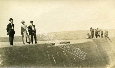 Sepia tone photo of the Heart River Dam with three people on the structure: Bertha Mann, Chris Kring (?) and unknown. 1911.