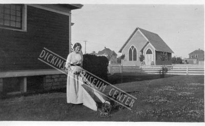 Photo of Ruth Maser mowing the side yard of the Maser Home in Dickinson. In the background is the St John Evangelical Lutheran Church. 1910-11.
